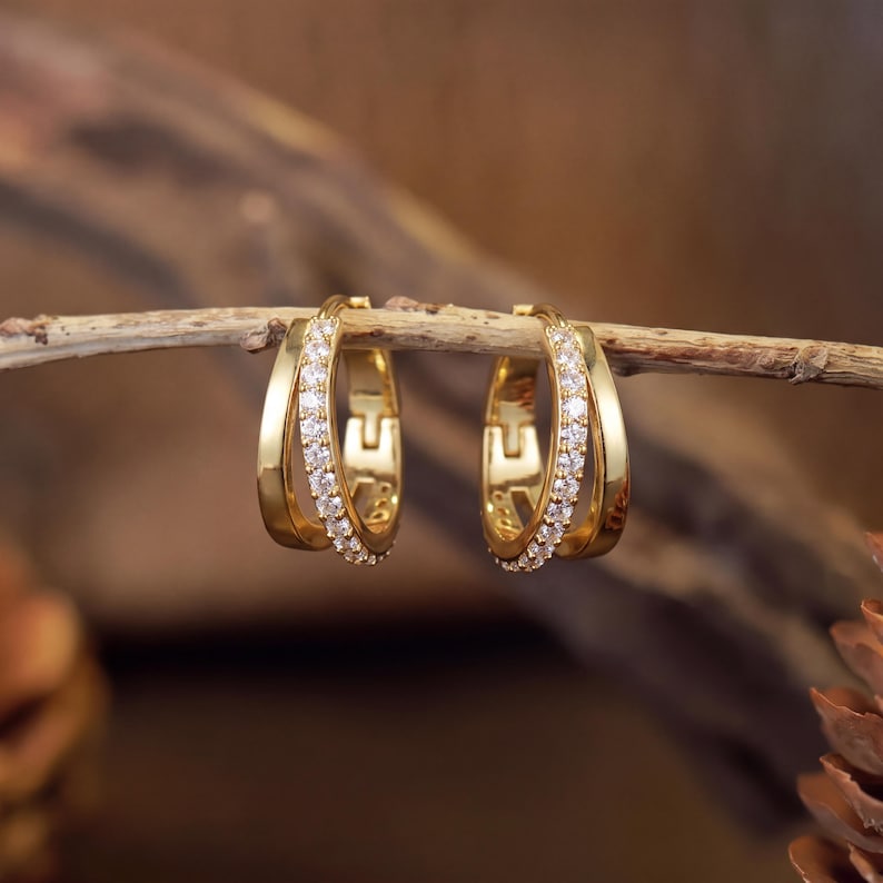May include: A pair of gold hoop earrings featuring a double-band design. One band is smooth and polished gold, while the other is encrusted with a row of small, clear, sparkling stones. The earrings are hinged and have a secure clasp mechanism.