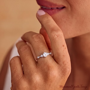 Può includere: Un anello color argento con una pietra centrale rotonda trasparente affiancata da due pietre più piccole su ciascun lato, montato su una sottile fascia metallica. Il testo MinimalGents Forge è visibile nell'angolo in basso a destra.