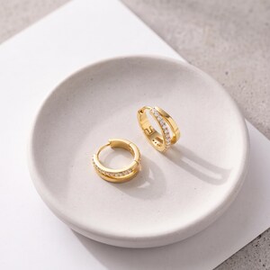 May include: A pair of gold hoop earrings featuring a double-band design. One band is smooth polished gold, while the other is encrusted with a row of small, clear, sparkling stones. These hinged huggie-style earrings have a secure clasp closure.