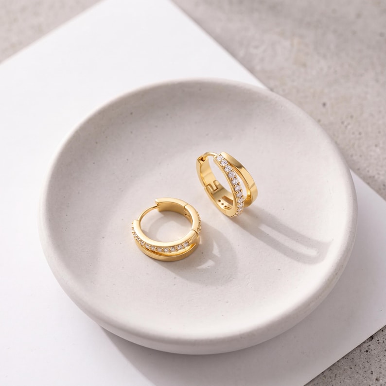 May include: A pair of gold hoop earrings featuring a double-band design. One band is smooth polished gold, while the other is encrusted with a row of small, clear, sparkling stones. These hinged huggie-style earrings have a secure clasp closure.