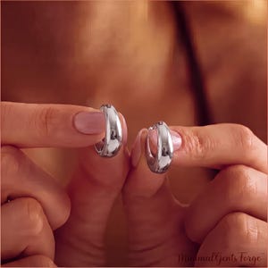 May include: A pair of small, polished silver hoop earrings with a thick, rounded profile. The earrings have a smooth, reflective surface and a classic circular shape. The text MinimalGentsForge is visible in the bottom right corner of the image.