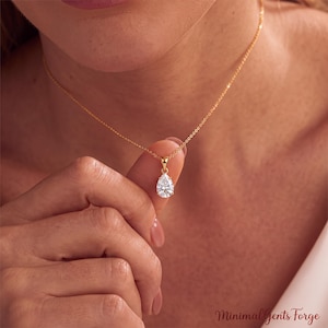 Puede incluir: Un collar de cadena dorada con un colgante de piedra preciosa transparente en forma de pera. El colgante cuelga de una delicada cadena de eslabones dorados que descansa sobre la clavícula. El texto Minimal Gents Forge aparece abajo a la derecha.