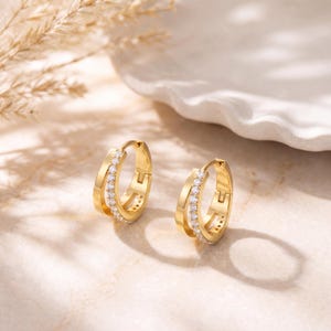 May include: A pair of gold hoop earrings featuring a double-band design. Each earring consists of a smooth outer band and an inner band encrusted with a row of small, clear, round-cut stones. The earrings have a hinged closure mechanism.