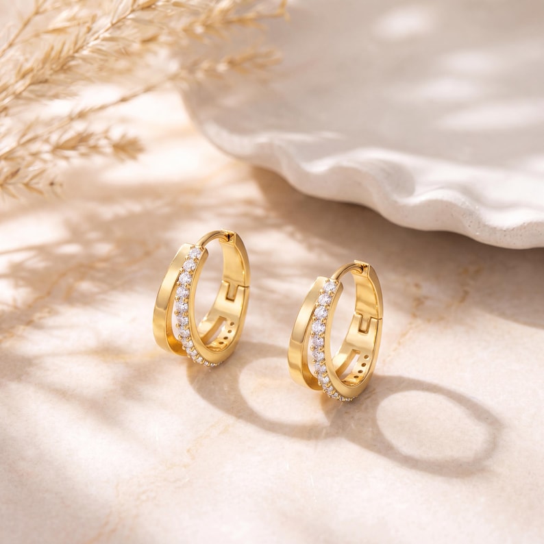May include: A pair of gold hoop earrings featuring a double-band design. Each earring consists of a smooth outer band and an inner band encrusted with a row of small, clear, round-cut stones. The earrings have a hinged closure mechanism.