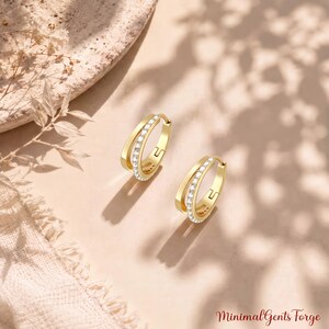 May include: A pair of gold hoop earrings featuring a double-band design with one band encrusted with small clear stones. The earrings are displayed with the text MinimalGentsForge in the bottom right corner, showcasing a polished finish.