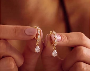 May include: A pair of gold hoop earrings featuring a row of small marquise-cut clear stones along the hoop and a single pear-shaped clear stone drop pendant. The earrings are held up to display the metal finish and the clear stone details.
