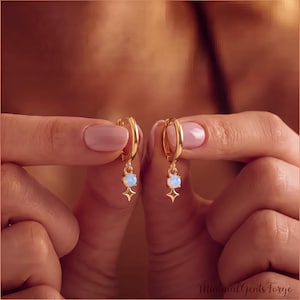 May include: A pair of gold hoop earrings featuring a small, round, iridescent opal stone suspended from each hoop. Below each opal is a four-pointed star charm. The earrings are crafted from a polished gold-tone metal with a bright finish.