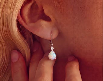 May include: A silver-toned leverback earring featuring a teardrop-shaped opal gemstone suspended beneath a small, round, clear faceted stone. The opal displays iridescent flashes of color, and the earring is secured to the earlobe.