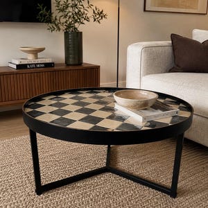 May include: A round coffee table with a black metal frame and a tabletop featuring a checkered pattern of alternating black and beige square tiles. A beige ceramic bowl sits on top of a book titled This Is Home placed on the table surface.