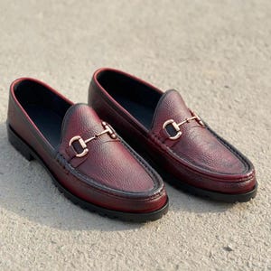 May include: A pair of deep burgundy loafers featuring a textured leather finish, a black trim along the edges, and a gold-toned metal horsebit detail across the vamp. The shoes are designed with a thick, black lugged rubber sole for traction.