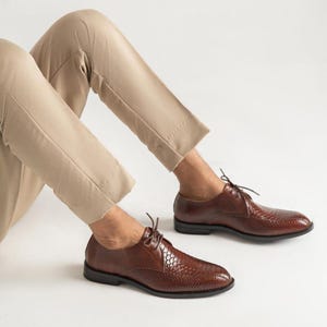 Op de afbeelding: Een paar bruine leren veterschoenen met een gestructureerd slangenleerachtig patroon op de zijkanten en de neus, gecombineerd met een beige broek. De schoenen hebben dunne bruine veters, een lage hak en een donkere zool voor een klassieke uitstraling.
