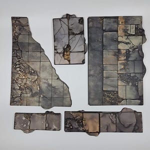 May include: A set of five flat, irregularly shaped terrain tiles featuring a dark, weathered stone floor pattern with cracked textures, visible grout lines, and patches of debris in shades of grey, brown, and muted gold, arranged on a plain surface.