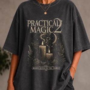 May include: A charcoal gray short-sleeve t-shirt featuring a graphic print with the text "PRACTICAL MAGIC 2" and "MAGIC RUNS IN THE FAMILY". The design includes two lit candles, a crescent moon, a cat silhouette, roses, and moths in gold tones.