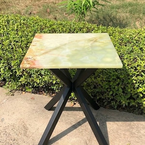 Puede incluir: Una mesa auxiliar cuadrada con un tablero de piedra ónix pulida en tonos verde claro y blanco con vetas naturales y una base de metal negro mate compuesta por cuatro patas anguladas que se cruzan en el centro para dar soporte.