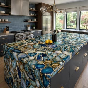 Puede incluir: Una isla de cocina con una encimera hecha de losas de piedra de ágata azul pulida con tonos variados de azul, blanco y dorado. La isla tiene gabinetes gris oscuro con manijas doradas y un porta utensilios negro con herramientas de cocina.
