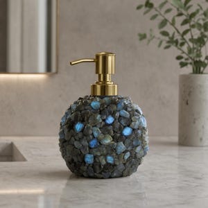 May include: A spherical soap dispenser featuring a textured exterior composed of dark grey and iridescent blue stone fragments. The dispenser is topped with a polished gold-toned metal pump mechanism, providing a functional contrast to the stone body.