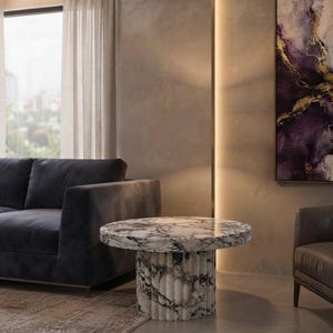 May include: A round coffee table featuring a white marble top with prominent dark grey veining and a cylindrical, fluted base made of the same white and grey marble. The table is positioned in a room with a dark grey sofa and a wall painting.