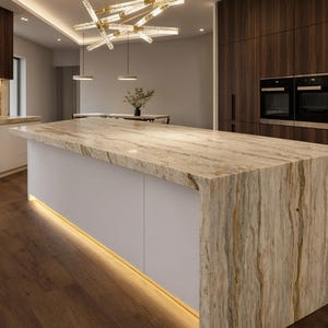 Puede incluir: Un interior de cocina con una gran isla central con encimera de piedra veteada en tonos beige y marrón con borde en cascada. La isla tiene gabinetes blancos con iluminación inferior, armarios de pared de madera oscura y una lámpara moderna.
