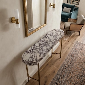Op de afbeelding: Een consoletafel met een langwerpig, ovaal blad van wit marmer met opvallende paarse aders en een gecanneleerde rand. De tafel wordt ondersteund door een slank, goudkleurig metalen frame met vier dunne poten en een centrale dwarsbalk.