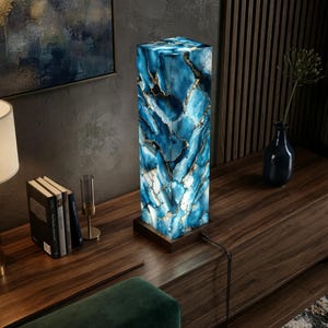 May include: A tall, rectangular table lamp featuring a translucent blue and white stone-like body with gold-toned veining. The lamp is mounted on a dark brown square wooden base and includes a black power cord extending from the side.