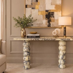 Op de afbeelding: Een consoletafel met een wit en grijs marmeren blad en twee poten van gestapelde bollen. Op de tafel staan een gouden vaas met groen blad, een gouden schaal, twee boeken Kinfolk en Louis Vuitton, twee witte ringvormige sculpturen en een lamp.