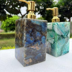 Può includere: Due dispenser di sapone rettangolari con corpo in pietra lucida e meccanismi a pompa in metallo dorato. Il dispenser anteriore presenta un motivo in pietra marmorizzata blu scuro e grigio, mentre quello posteriore mostra una pietra verde e bianca.