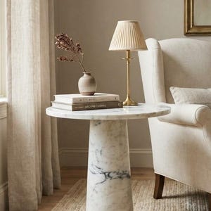 May include: A round white marble side table with a conical pedestal base. On the table sits a small ceramic vase with dried flowers, two stacked books, and a brass table lamp featuring a pleated beige fabric lampshade and a slender metal stem.