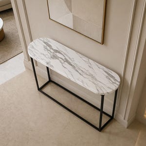 May include: A console table featuring an elongated oval top with a white and grey marble pattern. The table is supported by a thin, rectangular black metal frame base that provides a stable structure for the smooth, polished stone surface.