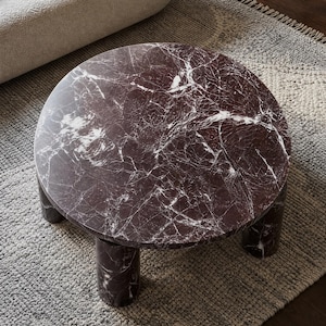 May include: A round coffee table crafted from deep burgundy marble with prominent white veining. The table features a smooth, circular tabletop supported by three thick, cylindrical legs, all finished in the same polished dark stone material.