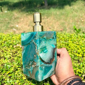 Puede incluir: Un dispensador de jabón cuadrado que presenta un mosaico de rodajas de piedra ágata en tonos verde azulado y verde con bandas naturales y texturas cristalinas. El dispensador está rematado con un mecanismo de bomba de tono dorado metálico.
