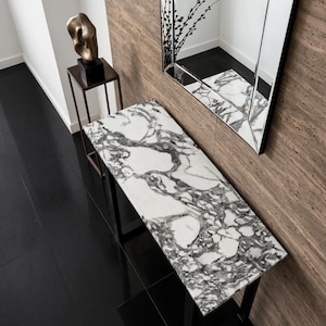 Op de afbeelding: Een rechthoekige sidetable met een wit marmeren blad met opvallende donkergrijze aders, ondersteund door een zwart metalen frame. De tafel staat tegen een getextureerde bruine muur, vergezeld door een hoge, dunne metalen standaard met een bronzen sculptuur.