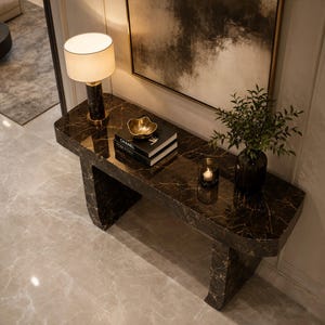 Puede incluir: Una mesa consola de mármol marrón oscuro con vetas blancas, que presenta una lámpara cilíndrica con pantalla blanca, dos libros apilados titulados Chanel y Tom Ford, un cuenco dorado, un pequeño portavelas de cristal y una planta verde en un jarrón oscuro.