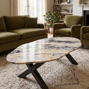 May include: An oval coffee table featuring a polished, multi-coloured marble top with veins of white, cream, black, and gold. The table is supported by a dark, matte black metal base with crossed legs that provide a stable foundation for the surface.