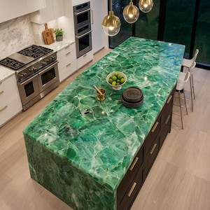 Puede incluir: Una gran isla de cocina con una encimera de piedra preciosa verde vibrante y translúcida con acabado pulido. La base de la isla es de madera oscura con herrajes dorados, con un cuenco blanco de limas, un mortero dorado y platos oscuros encima.