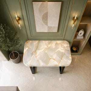 May include: A rectangular console table with a polished white and gold-veined stone top and black metal legs. The table is positioned against a sage green paneled wall featuring gold trim, two wall sconces, and a framed abstract beige wall art piece.