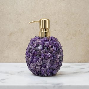 May include: A soap dispenser featuring a rounded body covered in small, textured purple amethyst stone chips. The top of the dispenser is equipped with a polished gold-toned metal pump mechanism designed for dispensing liquid soap or lotion.
