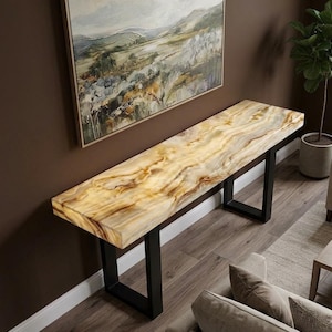May include: A long console table featuring a thick, rectangular top with a polished, translucent cream and brown marbled stone surface. The table is supported by two sturdy, black metal U-shaped legs that provide a stable base for the heavy stone top.