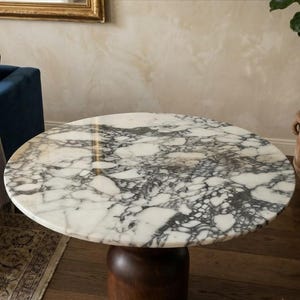 May include: A round table featuring a polished white marble top with prominent dark gray and black veining patterns. The table is supported by a smooth, cylindrical dark brown wooden base that extends downward from the center of the tabletop.