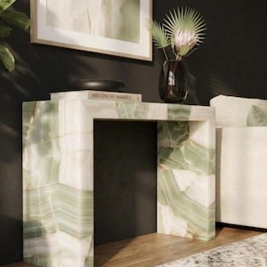 May include: A rectangular console table crafted from light green and white banded stone with a smooth finish. A book titled Mid-Century Marvels and a dark glass vase containing a palm leaf and a flower are placed on the table surface.
