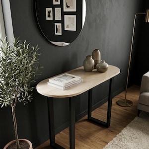 Op de afbeelding: Een consoletafel met een ovaal blad van licht hout en zwarte, U-vormige metalen poten. Drie keramische vazen in verschillende tinten beige en bruin staan op het oppervlak naast twee gestapelde boeken met witte covers en zwarte tekst op de rug.