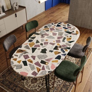 Op de afbeelding: Een ovale eettafel met een wit oppervlak voorzien van een veelkleurig terrazzopatroon van roze, gele, blauwe en grijze steenfragmenten. De tafel heeft dunne zwarte metalen poten en is omringd door vier stoelen met groene en grijze stoffen bekleding.
