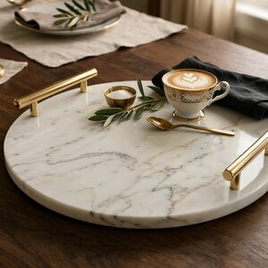 May include: A round white marble serving tray featuring natural grey veining and two polished gold-tone metal handles attached to the sides. The tray is smooth and circular, providing a flat surface for serving items like a cup, spoon, and small bowl.