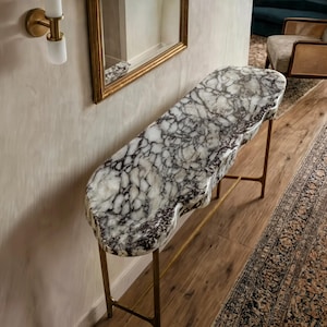 May include: A console table featuring an elongated, organic-shaped top made of white and black veined marble. The tabletop has a thick, polished edge and is supported by a slender, metallic gold-toned frame with four thin legs and a horizontal support bar.