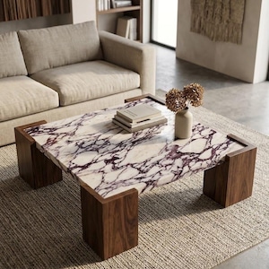 May include: A square coffee table featuring a white marble top with prominent purple veining, supported by four thick, dark brown wooden block legs. A small beige vase with dried flowers and a stack of two books sit on the marble surface.