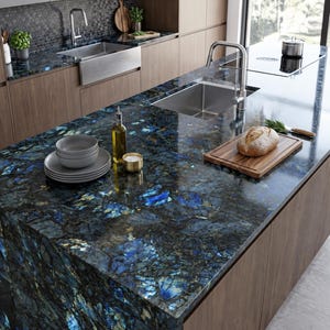 Puede incluir: Una isla de cocina con encimera de piedra labradorita azul pulida con borde en cascada, fregadero de acero inoxidable con grifo cromado, tabla de cortar de madera con un pan, una botella de aceite de vidrio y un juego de cuencos grises.