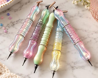 May include: Five diamond painting pens with contoured, translucent, and glittery bodies in pastel shades of pink, blue, green, and yellow. Each pen features a metallic tip, a T-shaped top, and a decorative silver-toned band around the center.
