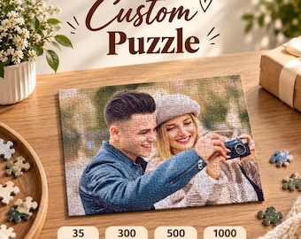 May include: A custom jigsaw puzzle featuring a photograph of a person in a denim jacket and a person in a grey beret holding a camera. The puzzle is displayed on a wooden surface with loose pieces and text reading Custom Puzzle and piece counts.