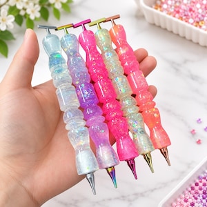 May include: Five diamond painting pens with ergonomic, multi-faceted resin handles in translucent blue, purple, hot pink, iridescent white, and coral orange. Each pen features a metallic T-shaped top and a pointed metal tip for placing small beads.