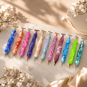 May include: A collection of eleven crafting pens featuring marbled, multi-coloured resin barrels with metallic accents. Each pen has a T-shaped top and a pointed metal tip, showcasing various colour combinations including blue, orange, pink, purple, and green.