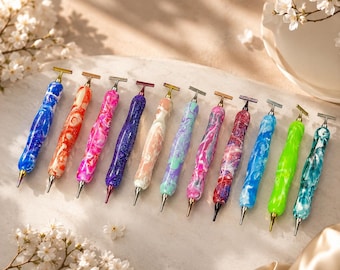 May include: A collection of eleven crafting pens with marbled, multi-colored resin barrels and metallic T-shaped tops. The pens feature various swirling color combinations including blue, orange, pink, purple, cream, teal, and bright green.